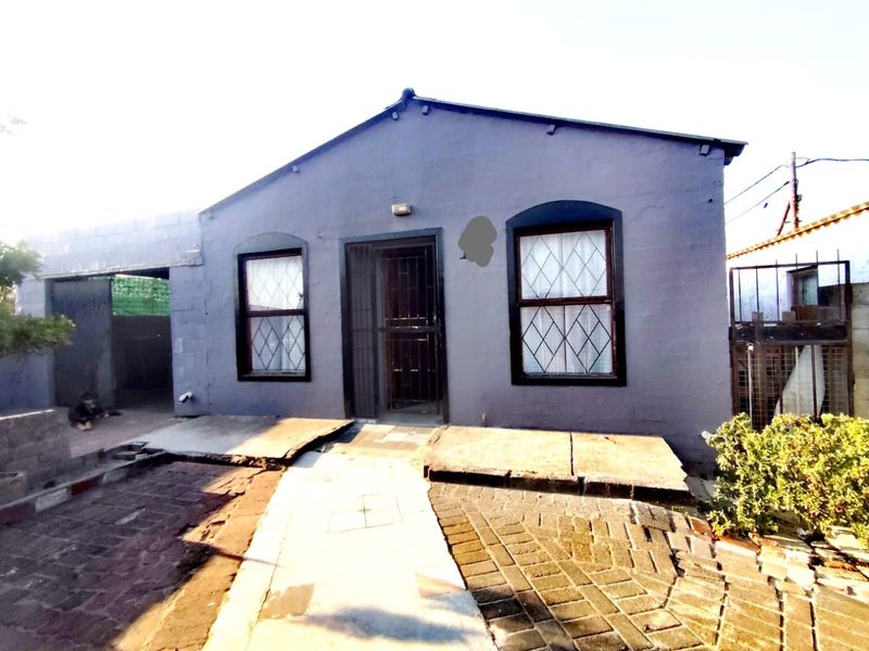 Front elevation of a single-story house featuring a paved entrance, security gate, and diamond-pattern windows, showing signs of wear on the exterior surfaces.