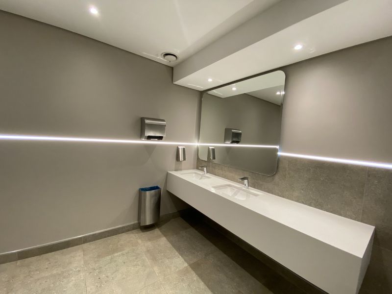 Modern commercial bathroom featuring a sleek double vanity with integrated sinks, large mirror, and ambient LED strip lighting.