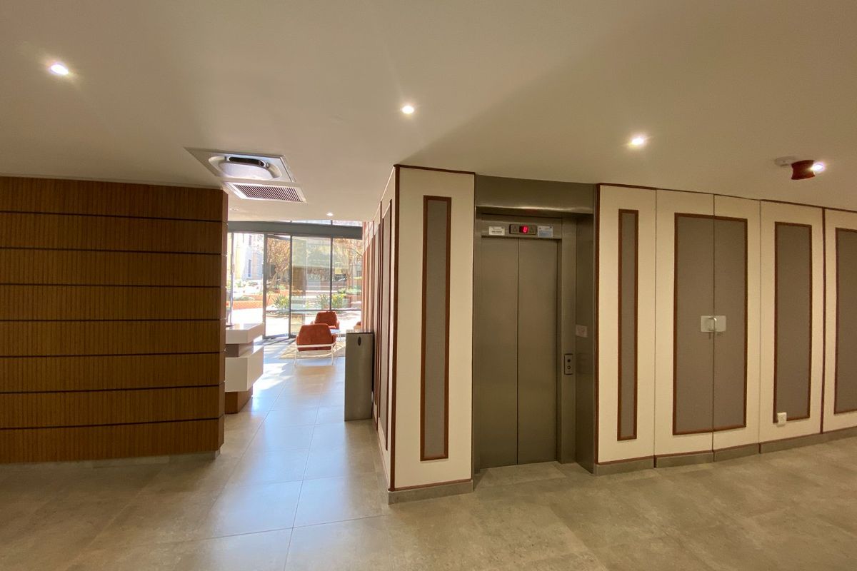 Modern commercial building lobby featuring a stainless steel elevator, tiled flooring, wood-paneled accents, and recessed lighting, leading to a bright entrance area.