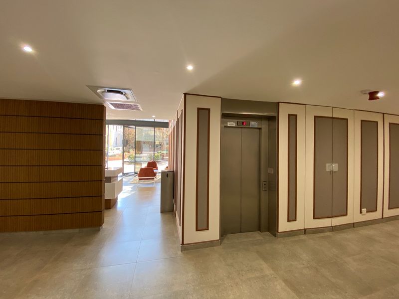 Modern commercial building lobby featuring a stainless steel elevator, tiled flooring, wood-paneled accents, and recessed lighting, leading to a bright entrance area.
