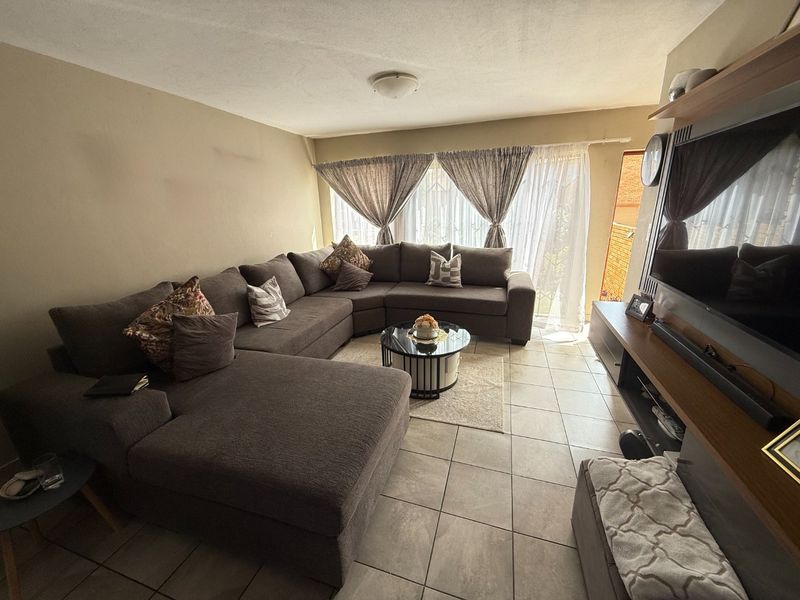A comfortable living room featuring tiled flooring and a built-in media shelving unit, with natural light entering through a large window with security gate.