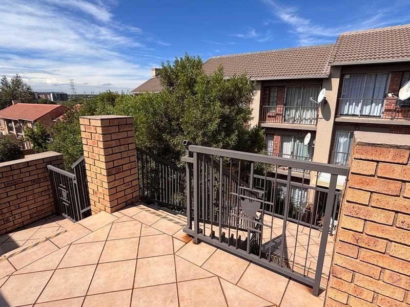 A spacious outdoor balcony featuring tiled flooring, brick pillars, and secure metal balustrades, overlooking the apartment complex grounds.