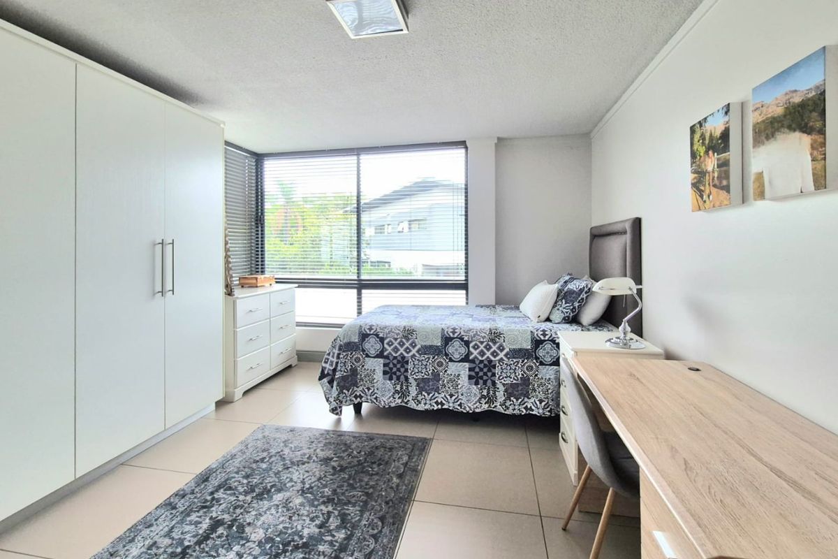 A well-lit bedroom featuring neutral tiled flooring, a large built-in wardrobe, and expansive windows with venetian blinds.