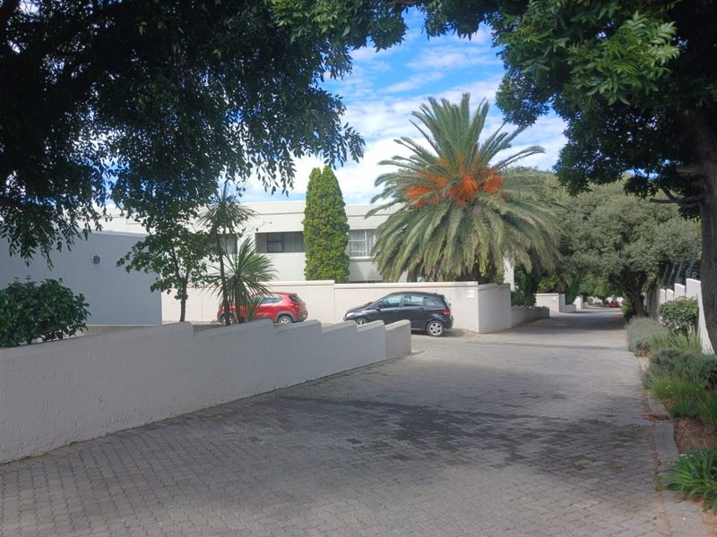 A view of the apartment complex entrance featuring a paved driveway, boundary walls with electric fencing, and established garden landscaping.