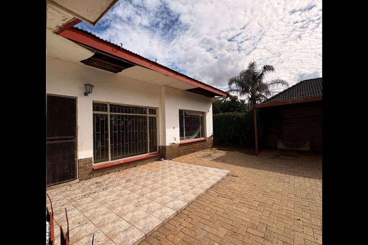 Rear exterior view of the property featuring a tiled patio, paved area, and security bars on windows, showing signs of significant wear and maintenance needs.