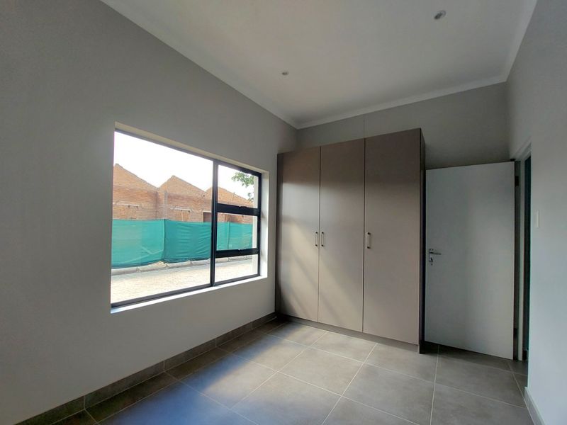 A clean, modern bedroom featuring neutral tiled flooring, built-in cupboards, and large aluminium-framed windows providing ample natural light.