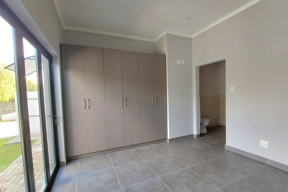 A bright, modern bedroom featuring neutral grey tones, large built-in cupboards, tiled flooring, and direct access to a private patio and en-suite bathroom.