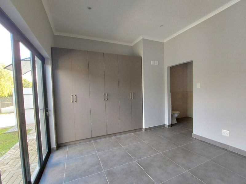 A bright, modern bedroom featuring neutral grey tones, large built-in cupboards, tiled flooring, and direct access to a private patio and en-suite bathroom.