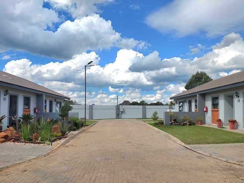 A view of the townhouse complex entrance featuring a paved driveway, well-maintained gardens, and modern exterior finishes under a clear blue sky.
