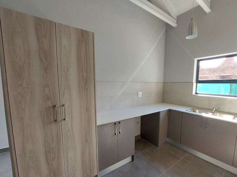 A modern, empty kitchen featuring light wood-tone cabinetry, neutral tiled backsplash, stainless steel sink, and exposed ceiling beams with pendant lighting.