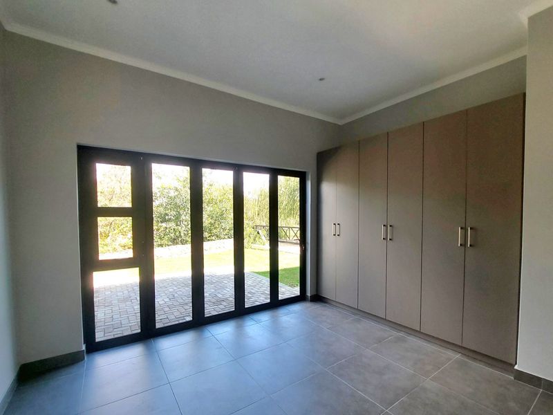 A bright, modern bedroom featuring neutral tiled flooring, extensive built-in cupboards, and large aluminium stacker doors opening onto a paved patio area.