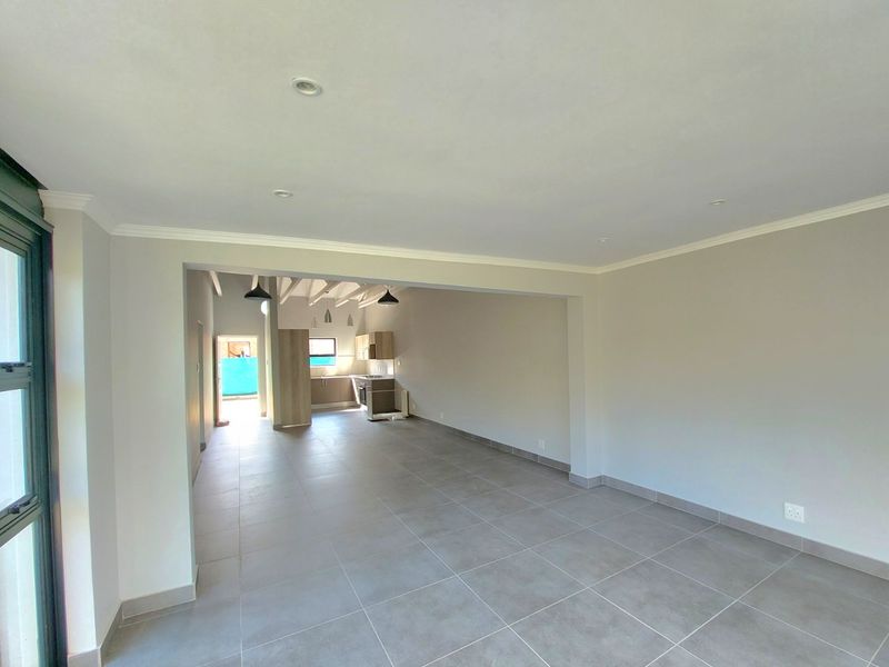 Spacious open-plan living area featuring neutral tiled flooring, recessed lighting, and a seamless transition into a functional kitchen space with exposed ceiling beams.