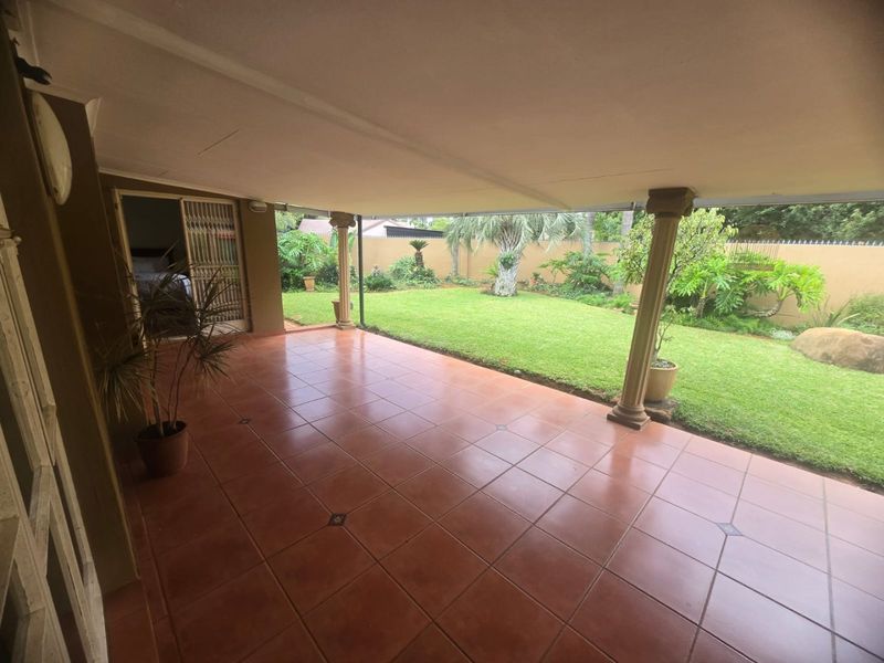 A spacious covered patio featuring tiled flooring and support columns, overlooking a well-maintained private garden with a perimeter boundary wall.