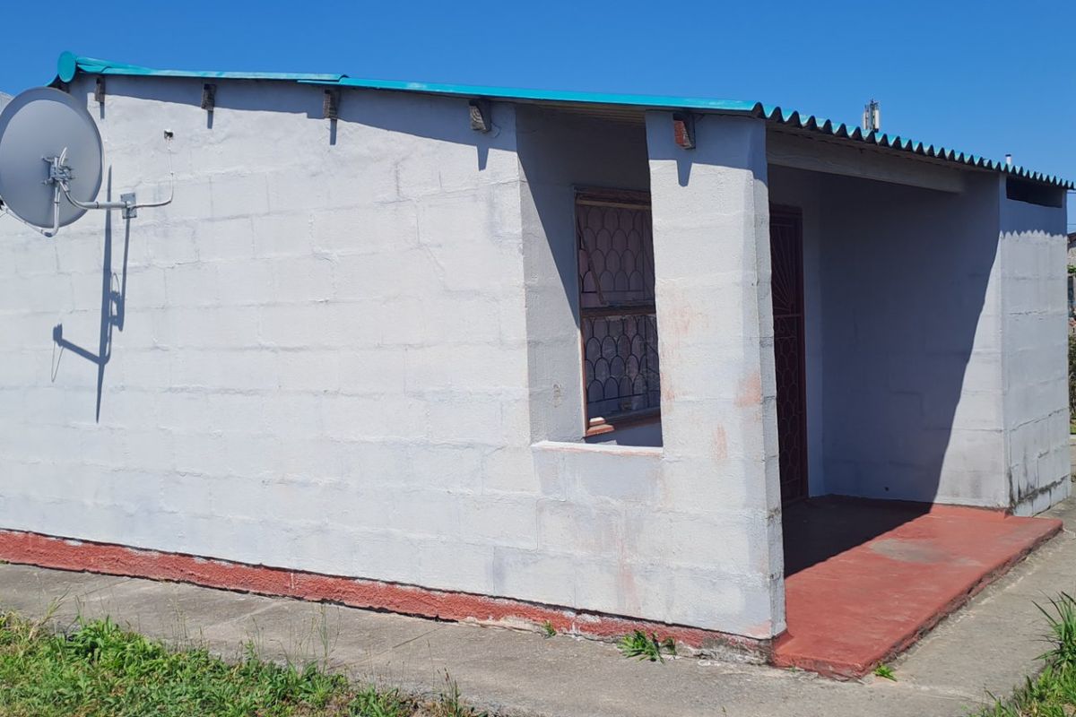 Rear view of a single-story concrete block house featuring a corrugated metal roof, a small covered patio area, and an attached satellite dish.