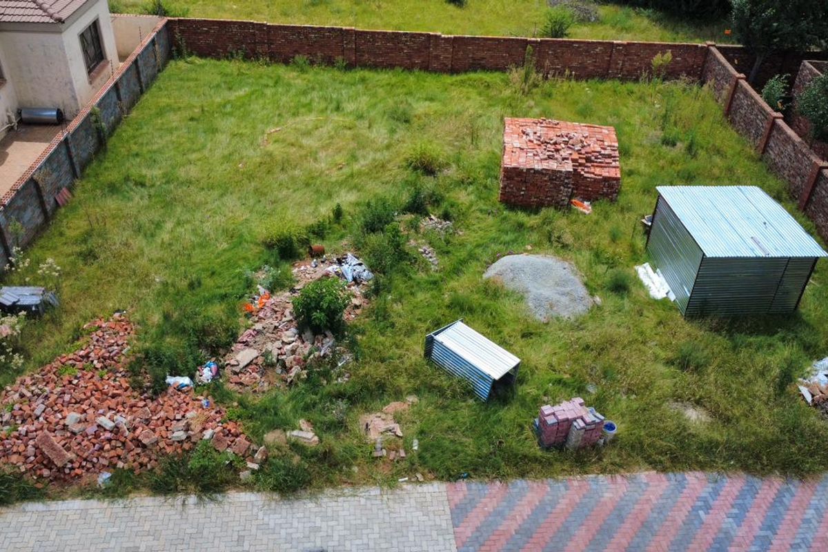 Aerial view of a vacant residential plot featuring a perimeter brick wall, paved driveway access, and scattered construction materials on site.