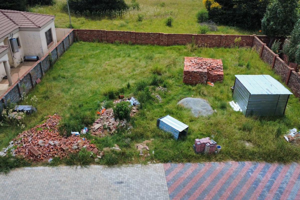 Aerial view of a vacant residential plot featuring a brick boundary wall, paved driveway, and scattered construction materials on overgrown grass.