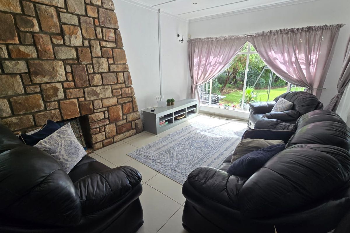 A living room featuring a prominent stone fireplace, tiled flooring, and large windows providing natural light and garden views.