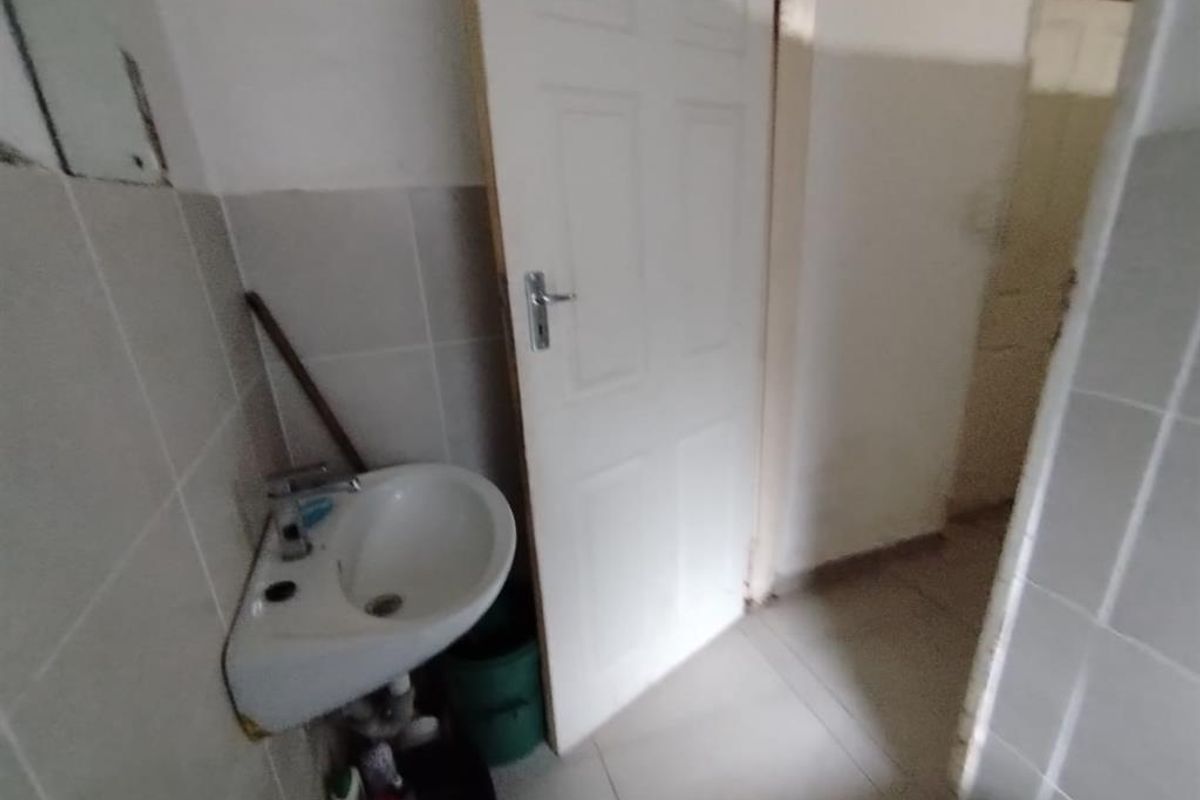 A small bathroom featuring a wall-mounted corner basin, tiled walls, and tiled flooring, currently in need of cosmetic updates and repairs.