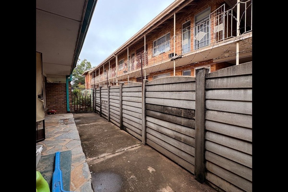 Exterior view of the apartment complex showing a paved walkway, concrete boundary wall, and multi-story brick facade with security bars.