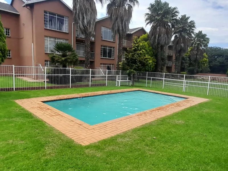 Communal swimming pool area featuring a brick-edged pool, well-maintained lawn, and secure metal perimeter fencing set against the townhouse complex facade.