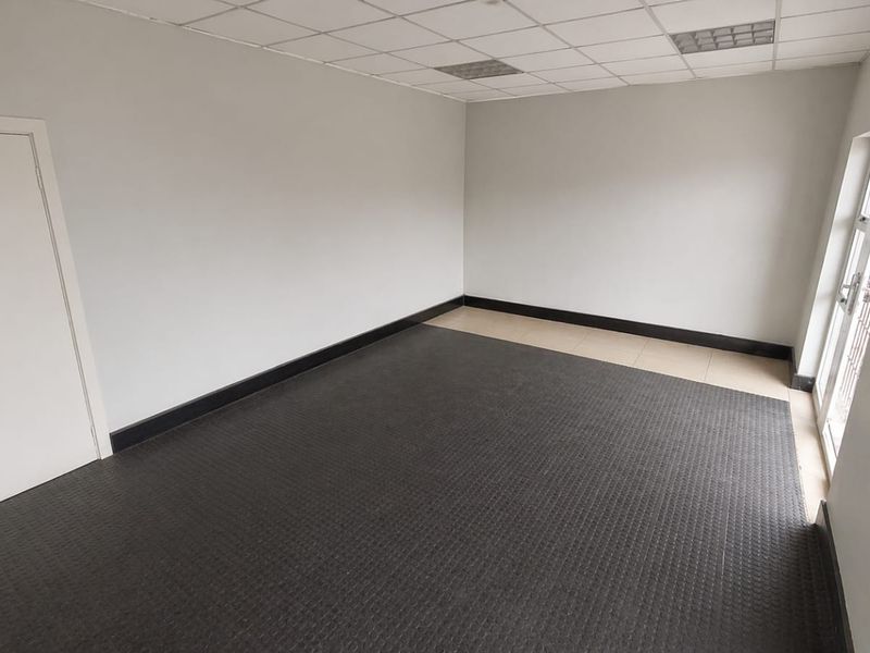 A clean, empty commercial office space featuring a suspended ceiling, recessed lighting, and durable rubber flooring with direct access to an exterior entrance.