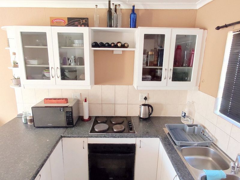 Compact kitchen featuring white cabinetry, dark laminate countertops, built-in electric hob and oven, and a stainless steel sink with tiled backsplash.