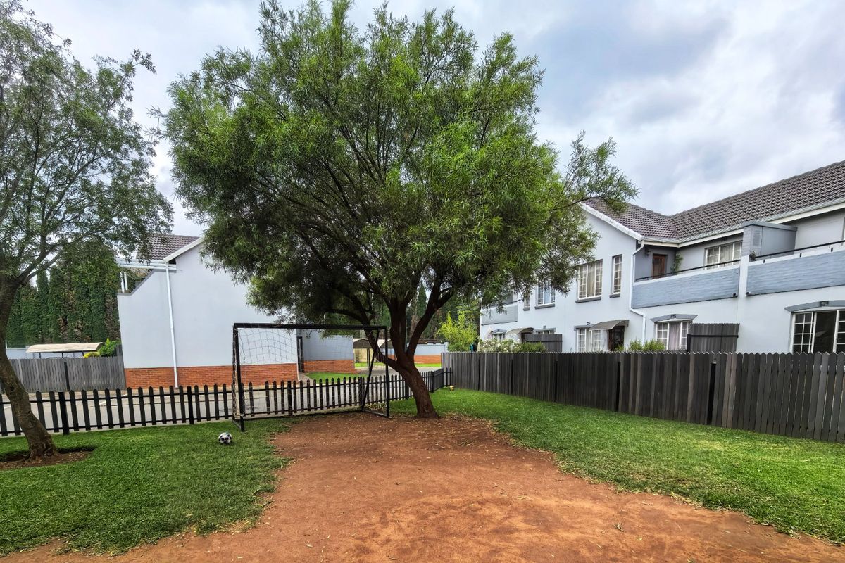 A communal garden area featuring a lawn, mature trees, and timber fencing, situated between apartment blocks with paved access.