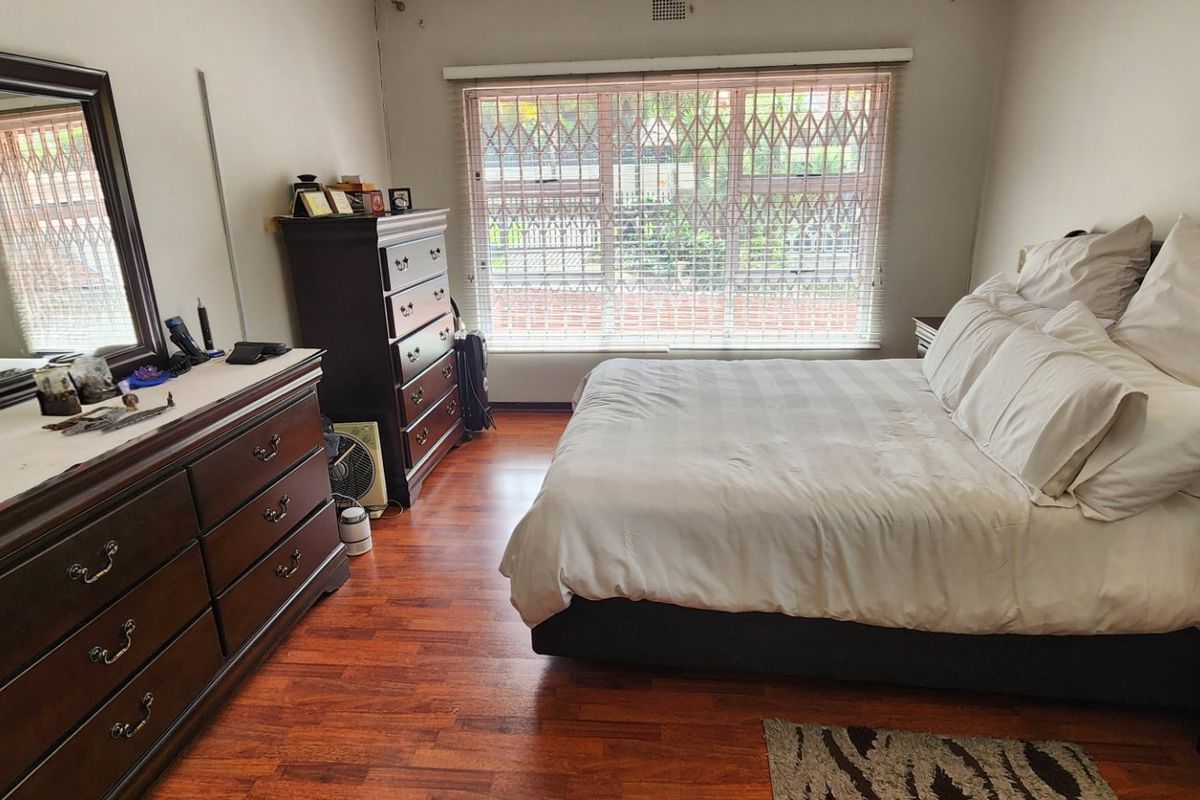 A spacious bedroom featuring warm-toned laminate flooring, a large window with security bars, and neutral wall finishes.