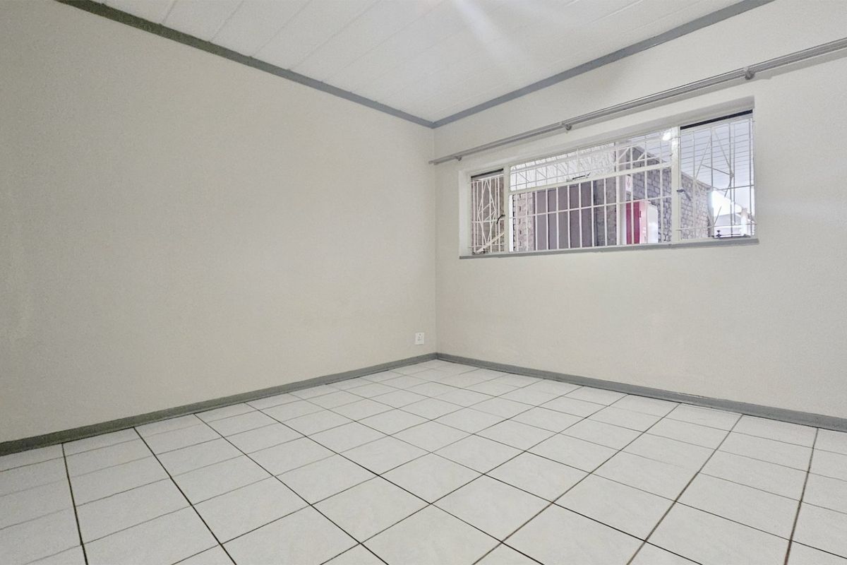 A clean, empty bedroom featuring white tiled flooring, built-in cupboards, and a window fitted with security bars.