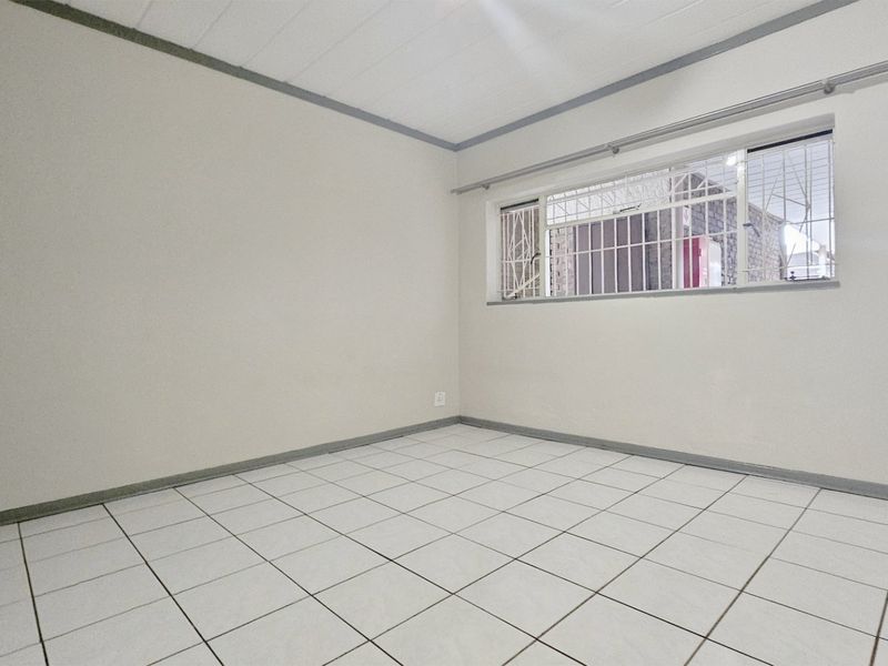 A clean, empty bedroom featuring white tiled flooring, built-in cupboards, and a window fitted with security bars.