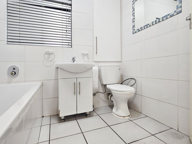 A clean, functional bathroom featuring a bathtub, vanity basin, toilet, and built-in storage with neutral white wall and floor tiling.