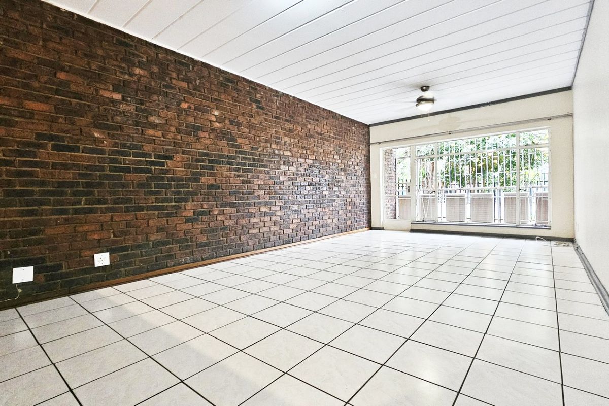 Spacious living area featuring a prominent exposed brick feature wall, neutral tiled flooring, and a large window with security gate access.