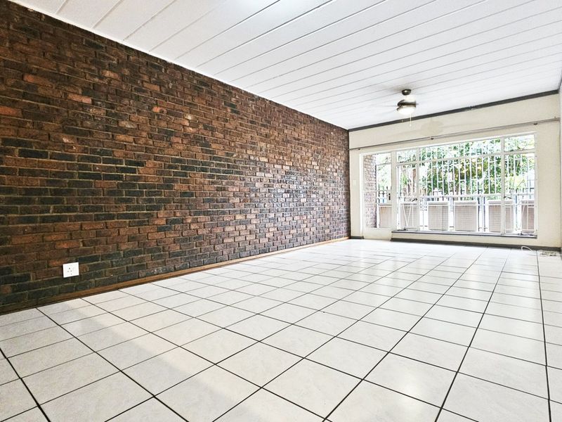 Spacious living area featuring a prominent exposed brick feature wall, neutral tiled flooring, and a large window with security gate access.