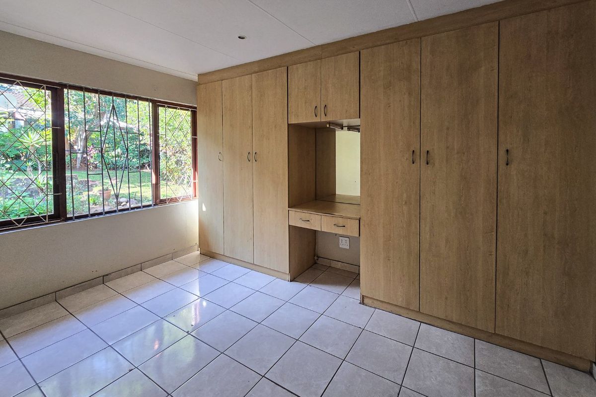 A bright, empty bedroom featuring extensive built-in wooden cupboards with an integrated vanity area, tiled flooring, and large windows with security bars.