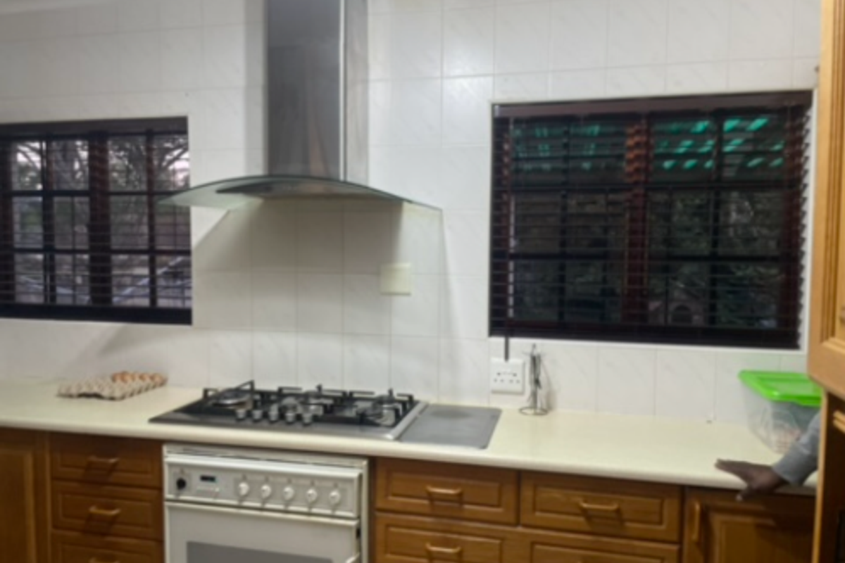 A functional kitchen featuring wooden cabinetry, a gas hob with an integrated extractor fan, and tiled splashbacks between two windows.