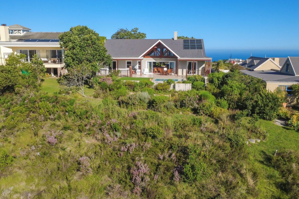 Aerial view of a residential property featuring a swimming pool, covered patio, and solar panels, set within a landscaped garden with coastal views.