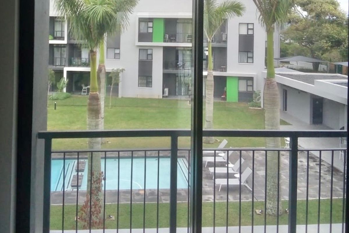 A balcony with tiled flooring and a metal balustrade, offering a view of the complex garden and swimming pool area.