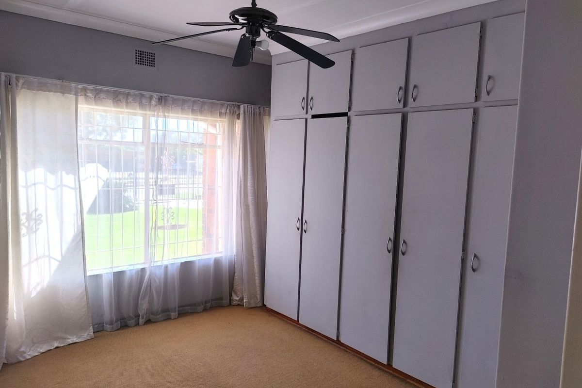 A bedroom featuring built-in floor-to-ceiling cabinetry, a ceiling fan, and large windows with security bars overlooking the garden.