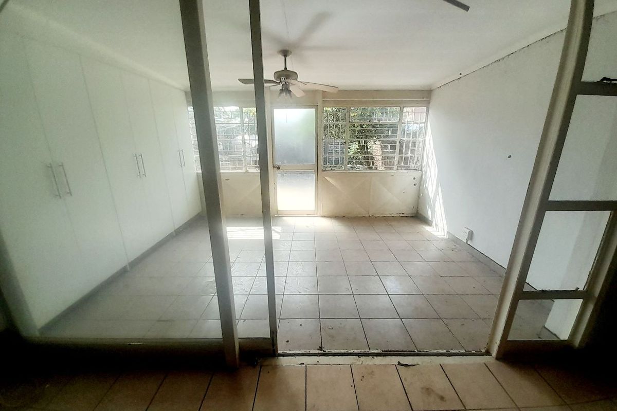An empty living area featuring tiled flooring, built-in cupboards, a ceiling fan, and large windows with security bars, offering a blank canvas for renovation.