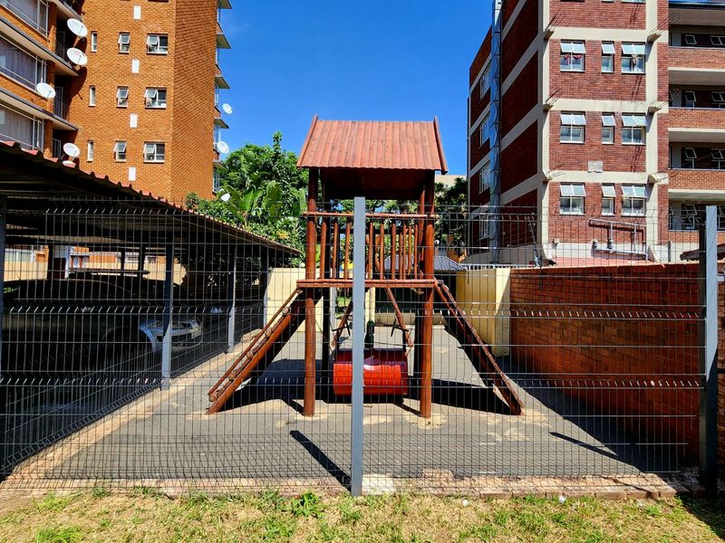 Outdoor communal playground area situated between apartment buildings, featuring fixed play equipment and secure perimeter fencing.