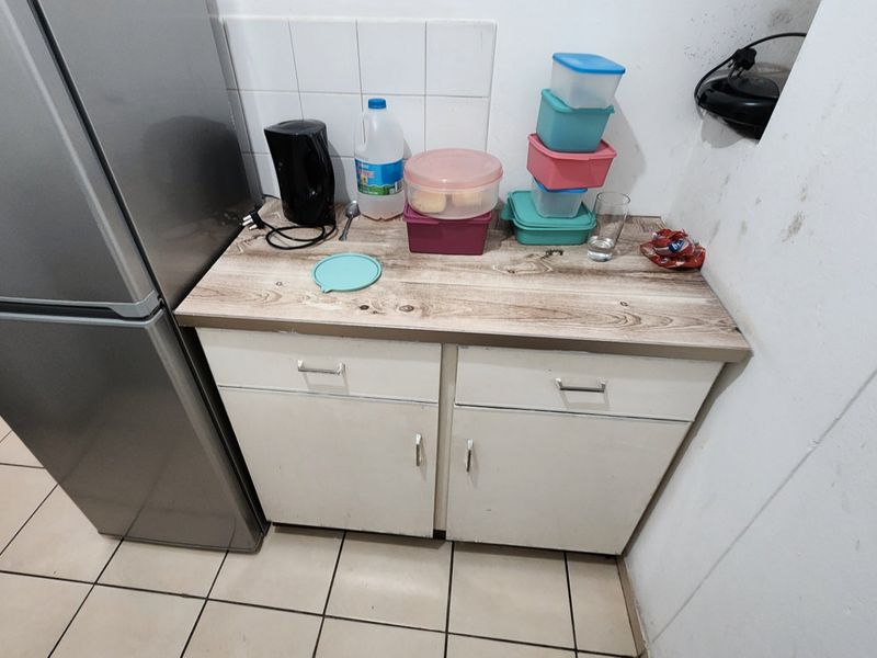 A compact kitchen area featuring laminate countertops, base cabinetry, and tiled splashbacks, showing signs of wear and wall staining.