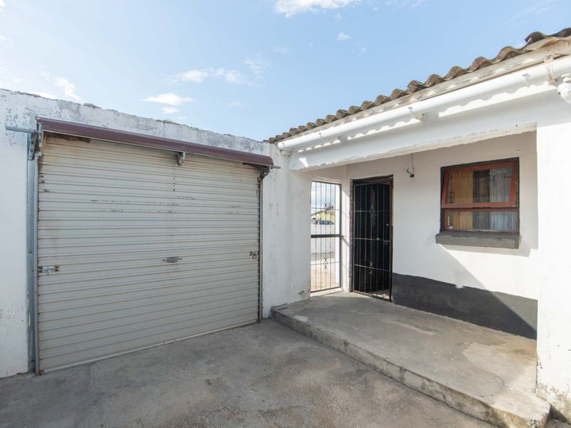 Rear exterior view showing a garage with a roller shutter door, a small concrete patio area, and security gate access to the property.