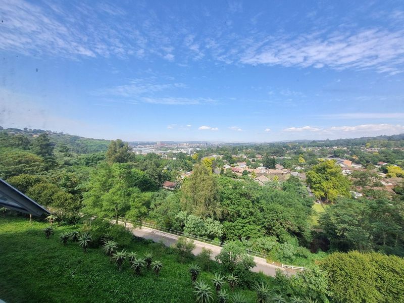 An elevated, scenic view overlooking a lush green suburban landscape with mature trees and distant residential areas under a clear blue sky.