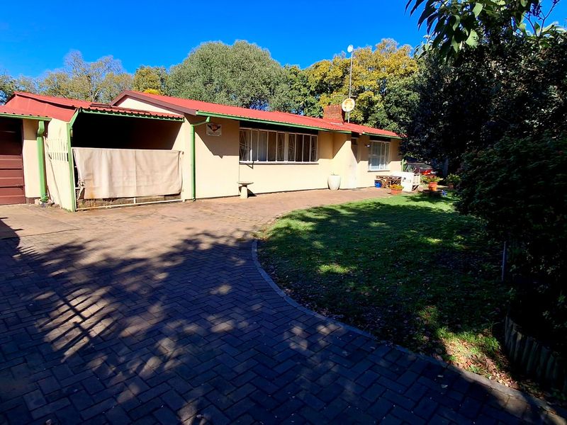 Rear view of the property featuring a paved driveway, covered patio area, and a spacious garden with mature trees.