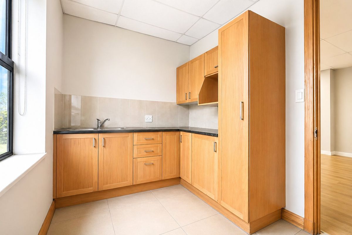 A functional kitchenette featuring light wood cabinetry, dark countertops, a stainless steel sink, and tiled flooring in a commercial office setting.