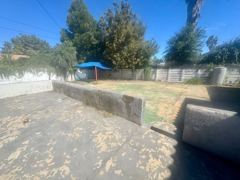 A rear outdoor area featuring a concrete patio with a retaining wall, a large garden space, and a boundary wall with a water tank.