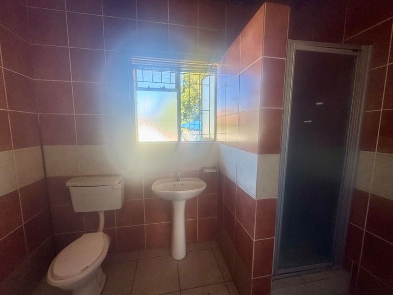 A dated bathroom featuring a pedestal basin, toilet, and shower enclosure with full-height wall tiling and a window with security bars.