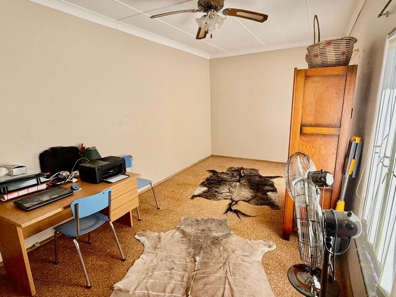 A bedroom featuring a ceiling fan, coving, and vertical blinds, currently utilized as a home office space with worn carpeting.