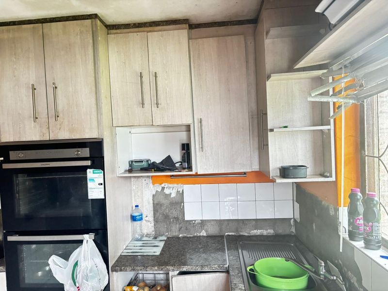 A kitchen area featuring built-in double ovens, laminate cabinetry, and a stainless steel sink, currently requiring wall repairs and finishing to the splashback area.