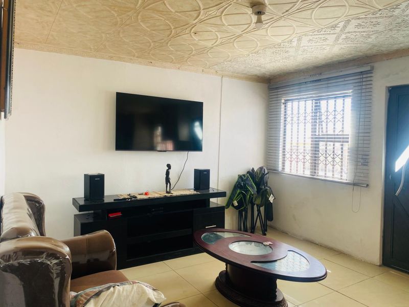 A living room featuring decorative ceiling tiles, tiled flooring, and a window fitted with security bars and venetian blinds.